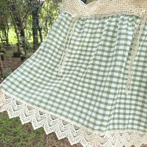 May include: A green and white gingham skirt with cream-coloured lace trim along the bottom edge. The skirt has a cream-coloured crocheted waistband and cream-coloured ties. The skirt is made of a lightweight fabric.