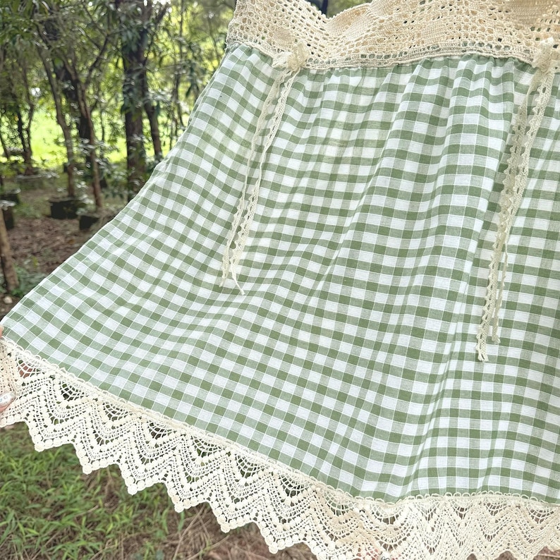 May include: A green and white gingham skirt with cream-coloured lace trim along the bottom edge. The skirt has a cream-coloured crocheted waistband and cream-coloured ties. The skirt is made of a lightweight fabric.