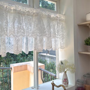 May include: A white lace window valance with a scalloped edge hangs above a window. The valance is made of delicate, detailed lace. The window looks out onto a green, leafy outdoor scene. A vase of white flowers sits on the windowsill.