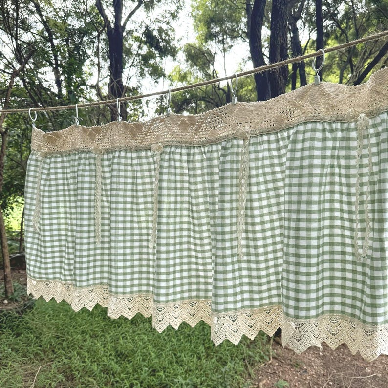May include: A green and white gingham curtain with a beige lace trim. The curtain is suspended from metal rings on a rope, set against a backdrop of trees and foliage. The curtain has a rustic, country style.
