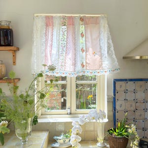以下が含まれることがあります： ピンク、白、花柄のパネルが付いたレースのカーテンが付いたキッチンの窓。窓からは庭が見えます。棚には瓶が置かれ、カウンターには花瓶、白い蘭、小さな鉢植えが置かれています。