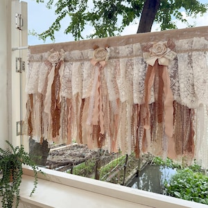 May include: A cream, tan, and brown lace window valance with fabric roses and ribbons. The valance has multiple layers of lace and fabric, creating a vintage and romantic look. This window treatment is designed to add a touch of elegance to any room, with a classic, timeless design.