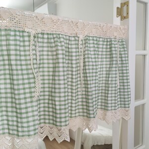 May include: A green and white gingham curtain with cream-coloured lace trim and top. The curtain hangs in a doorway, with lace trim at the top and bottom edges. The curtain is made of a lightweight fabric, suitable for a kitchen or bedroom.