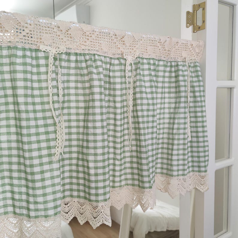 May include: A green and white gingham curtain with cream-coloured lace trim and top. The curtain hangs in a doorway, with lace trim at the top and bottom edges. The curtain is made of a lightweight fabric, suitable for a kitchen or bedroom.