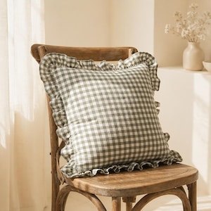 Peut inclure: Un coussin carré avec un motif à carreaux vichy vert sauge et blanc. Le coussin est agrémenté d'un bord en tissu volanté sur les quatre côtés, créant un cadre décoratif autour du motif central à carreaux carrés.