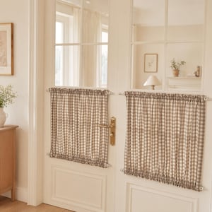 Puede incluir: Dos cortinas de cuadros vichy en beige y blanco montadas en una puerta doble blanca. Cada cortina está fruncida en una barra superior e inferior, cubriendo los paneles de cristal de la puerta. La tela presenta un pequeño patrón de cuadros.