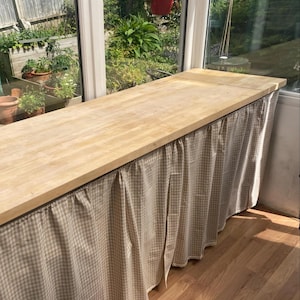 K&ouml;nnte beinhalten: Eine hellfarbene Holz-Arbeitsplatte mit einem beigefarbenen und wei&szlig; karierten Stoffrock. Die Arbeitsplatte befindet sich vor einem gro&szlig;en Fenster mit Blick auf einen Garten. Das Holz hat eine nat&uuml;rliche Maserung.
