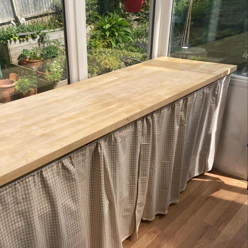 K&ouml;nnte beinhalten: Eine hellfarbene Holz-Arbeitsplatte mit einem beigefarbenen und wei&szlig; karierten Stoffrock. Die Arbeitsplatte befindet sich vor einem gro&szlig;en Fenster mit Blick auf einen Garten. Das Holz hat eine nat&uuml;rliche Maserung.