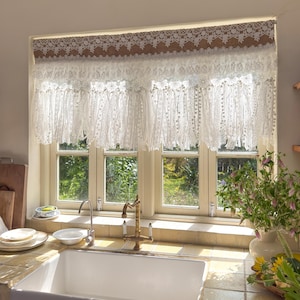 May include: A white lace window valance with a brown border. The valance is made of delicate, layered lace and hangs above a window with a view of a garden. The kitchen sink and faucet are visible below the window.