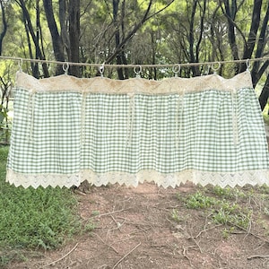 May include: A green and white gingham valance with cream lace trim. The valance is suspended from metal rings on a washing line. The fabric features a traditional check pattern, suitable for a country-style kitchen.