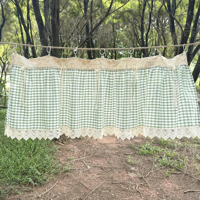 May include: A green and white gingham valance with cream lace trim. The valance is suspended from metal rings on a washing line. The fabric features a traditional check pattern, suitable for a country-style kitchen.