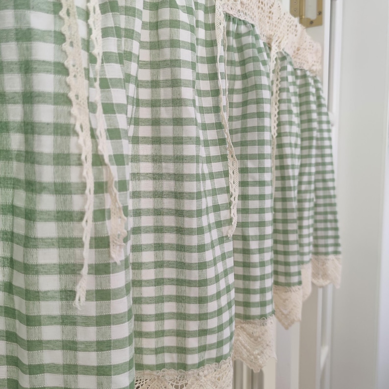 May include: Green and white gingham curtains with a cream lace trim. The curtains are made of a lightweight fabric and are hanging in front of a white door frame. The lace trim is a cream colour.