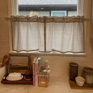 K&ouml;nnte beinhalten: Ein Fenster mit einem braun-wei&szlig; karierten Volant. Darunter befinden sich verschiedene K&uuml;chenartikel: ein gerahmtes Bild, eine Butterdose, Kochb&uuml;cher, ein Messerblock und Kanister.