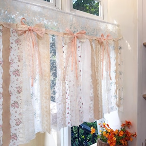May include: A decorative window valance in a vintage style. It features a mix of floral, lace, and burlap fabrics in white, cream, and beige. The valance is adorned with delicate pink bows, adding a touch of elegance to the window treatment.