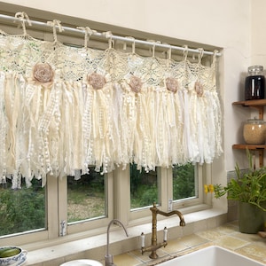 May include: Cream-colored lace window valance with fabric flower accents. The valance features layered lace and fabric strips, creating a textured, bohemian look. The valance is hung on a white rod above a window, adding a touch of vintage charm to the kitchen.