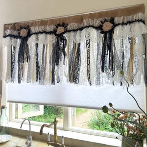 May include: A window valance featuring a tan burlap header adorned with three fabric rosettes and cascading strips of black, white, and cream lace, ribbon, and fabric trim. The valance hangs above a white pleated window shade in a kitchen setting.
