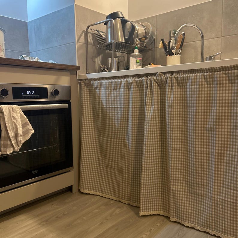 K&ouml;nnte beinhalten: K&uuml;cheninterieur mit Edelstahlbackofen und Sp&uuml;lbereich. Unter dem Waschbecken h&auml;ngt ein beige-wei&szlig; karierter Vorhang. Ein Geschirrkorb aus Metall h&auml;lt Geschirr und einen Wasserkocher. Ein Handtuch h&auml;ngt an der Backofent&uuml;r.