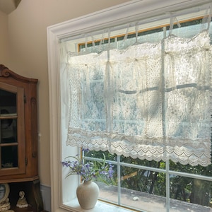 May include: A window with white lace curtains. The curtains have a scalloped edge and are tied with ribbons. A small vase with purple flowers sits on the windowsill. The window frame is white.