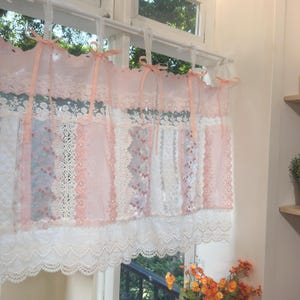 May include: A delicate window curtain with a layered design. The curtain features panels of white and pink lace, accented with peach-colored ribbons. The bottom edge has a scalloped lace trim, creating a romantic and vintage aesthetic. The curtain is hanging in front of a window.
