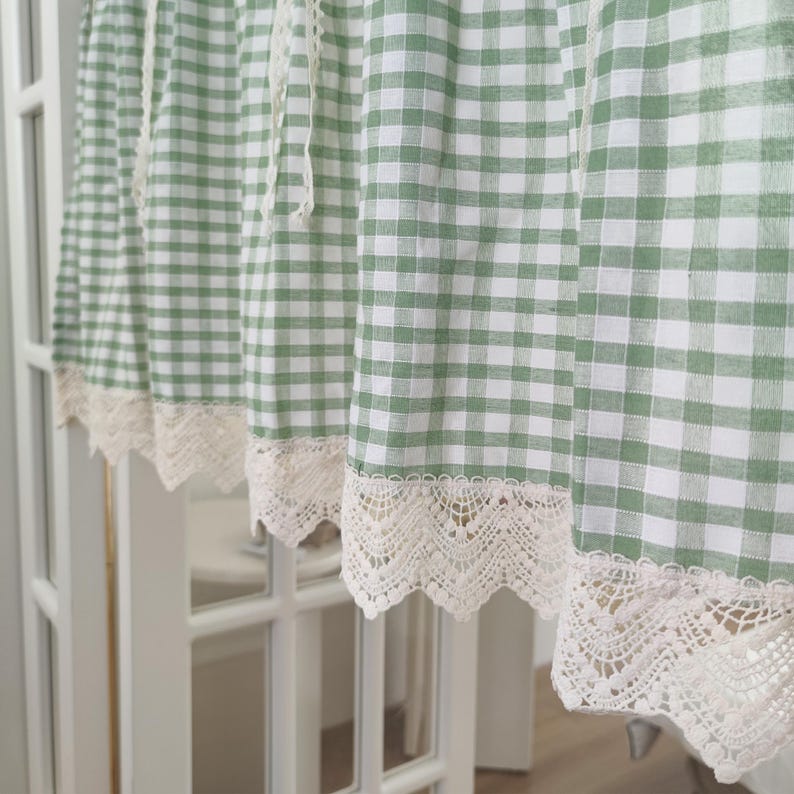 May include: A green and white gingham patterned curtain with a cream lace trim. The curtain hangs in front of a white door. The fabric has a classic checkered design, suitable for a kitchen or dining room.