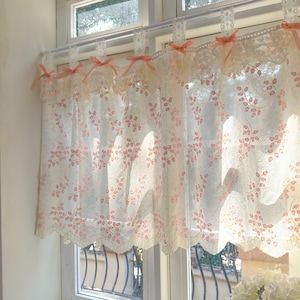 Könnte beinhalten: Transparente weiße Spitzen-Fenstervorhänge mit einem rosa Blumenmuster. Die Vorhänge sind mit einem gewellten Rand verziert und mit rosa Schleifen gebunden. Sie hängen an einer weißen Gardinenstange.