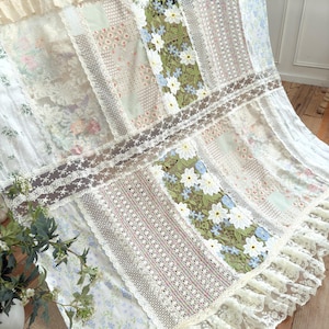 May include: A patchwork textile featuring various lace and floral patterns. The design incorporates white, cream, and pastel colors, with sections of embroidered flowers and delicate lace trim. The overall aesthetic is vintage and romantic.