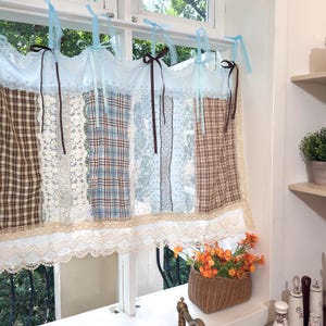 Può includere: Una tendina a patchwork per finestre con vari tessuti, tra cui scozzese, pizzo e motivi a pois. La tendina è nei toni del marrone, blu e bianco, con bordi in pizzo e nastri blu. Un cestino di vimini con fiori arancioni.