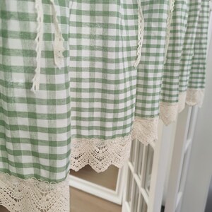 May include: Green and white gingham check curtains with cream lace trim. The curtains are hanging, showing a close-up of the fabric's texture. The design is a classic, country-style pattern, with a delicate lace border.