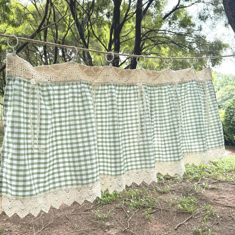 May include: Green and white gingham curtains with cream lace trim. The curtains are suspended from metal rings on a rope, set against a natural outdoor backdrop. The curtains have a country or farmhouse aesthetic.