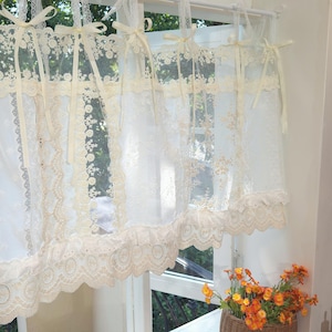 May include: Sheer white lace curtains with cream-colored trim and delicate floral patterns. The curtains are tied with cream-colored ribbons and hang from a white rod. A basket of orange flowers sits on the windowsill.