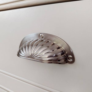 May include: A brushed nickel shell-shaped drawer pull is mounted on a cream-coloured cabinet door. The hardware features a detailed, fan-like design and is secured with two screws.