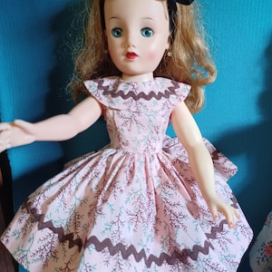 May include: A vintage doll wearing a pink dress with a brown and green floral pattern. The dress has a scalloped collar and a brown trim around the hem.