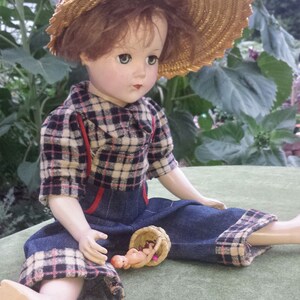 May include: A porcelain doll wearing a plaid shirt, blue denim overalls, and a straw hat. The doll is holding a small wicker basket with a baby doll inside.