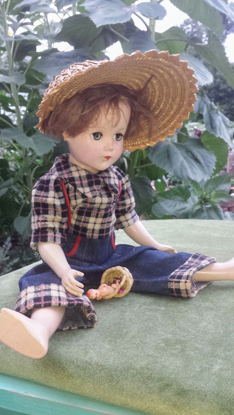May include: A porcelain doll wearing a plaid shirt, blue denim overalls, and a straw hat. The doll is holding a small wicker basket with a baby doll inside.
