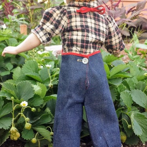 May include: A doll wearing a plaid shirt, denim overalls with a red trim, and a straw hat. The doll is standing in a garden with green plants and white flowers.