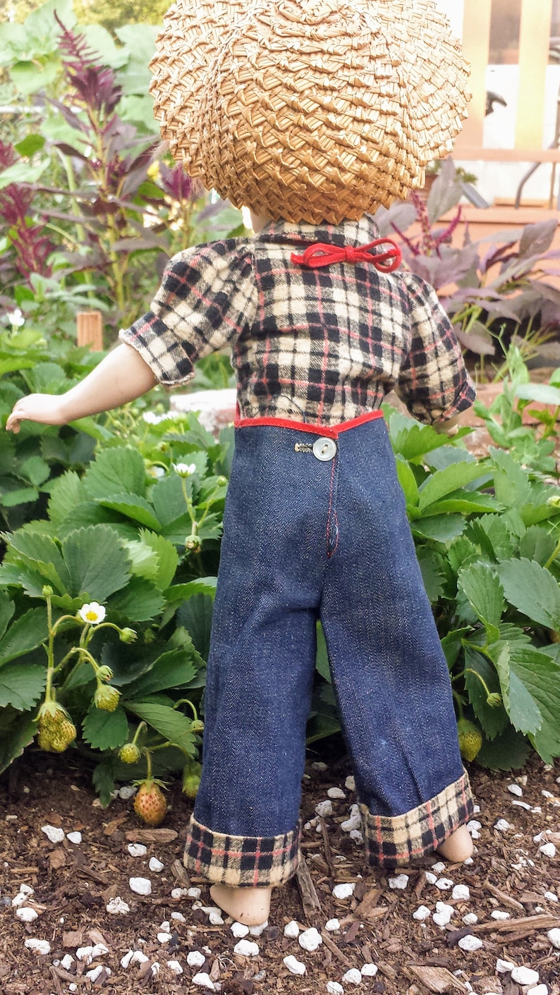 May include: A doll wearing a plaid shirt, denim overalls with a red trim, and a straw hat. The doll is standing in a garden with green plants and white flowers.