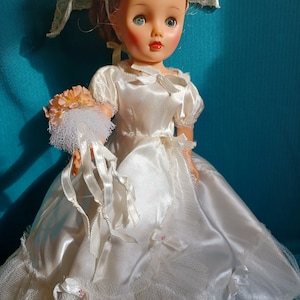 Könnte beinhalten: Eine Vintage-Brautpuppe in einem weißen Satin-Kleid mit einem Spitzen-Schleier und einem Strauß rosa Blumen.