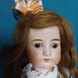 May include: Antique porcelain doll with brown hair and a peach-colored satin bow. The doll has painted brown eyes and rosy cheeks. She is wearing a white dress with lace trim.
