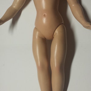 May include: A nude-colored plastic doll with long, straight hair. The doll is standing with its arms outstretched and its legs together. The doll has a smooth, shiny finish.