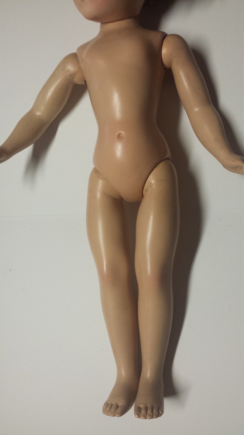 May include: A nude-colored plastic doll with long, straight hair. The doll is standing with its arms outstretched and its legs together. The doll has a smooth, shiny finish.
