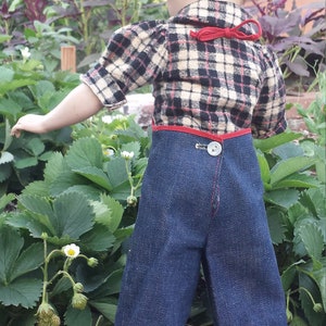 May include: A doll wearing a plaid shirt with a red ribbon and blue denim overalls with plaid trim.