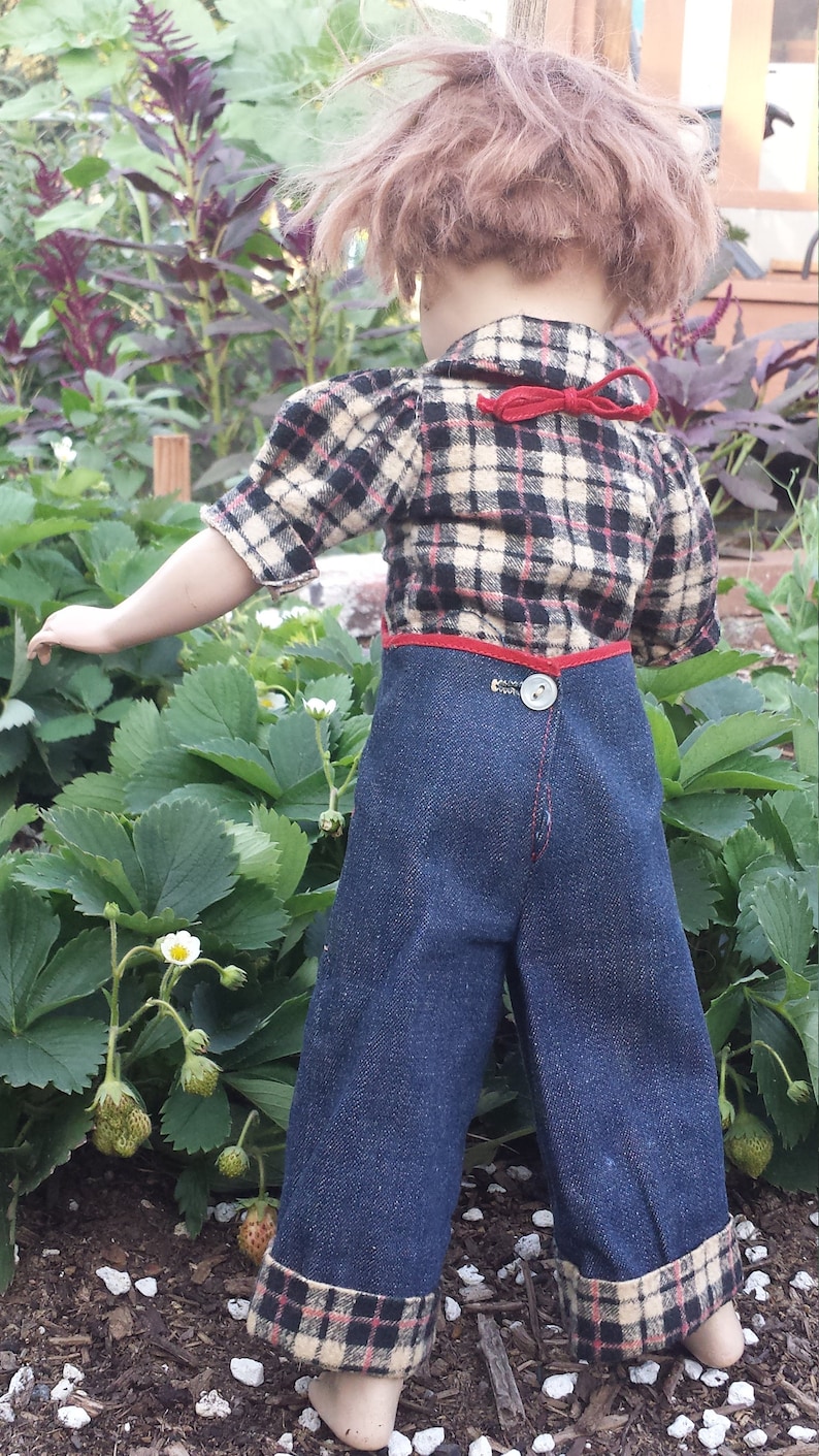 May include: A doll wearing a plaid shirt with a red ribbon and blue denim overalls with plaid trim.