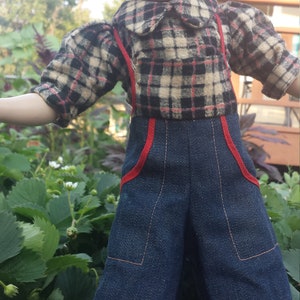 May include: A vintage doll with brown hair, wearing a plaid shirt and blue denim overalls with red trim. The doll is standing in a garden setting.