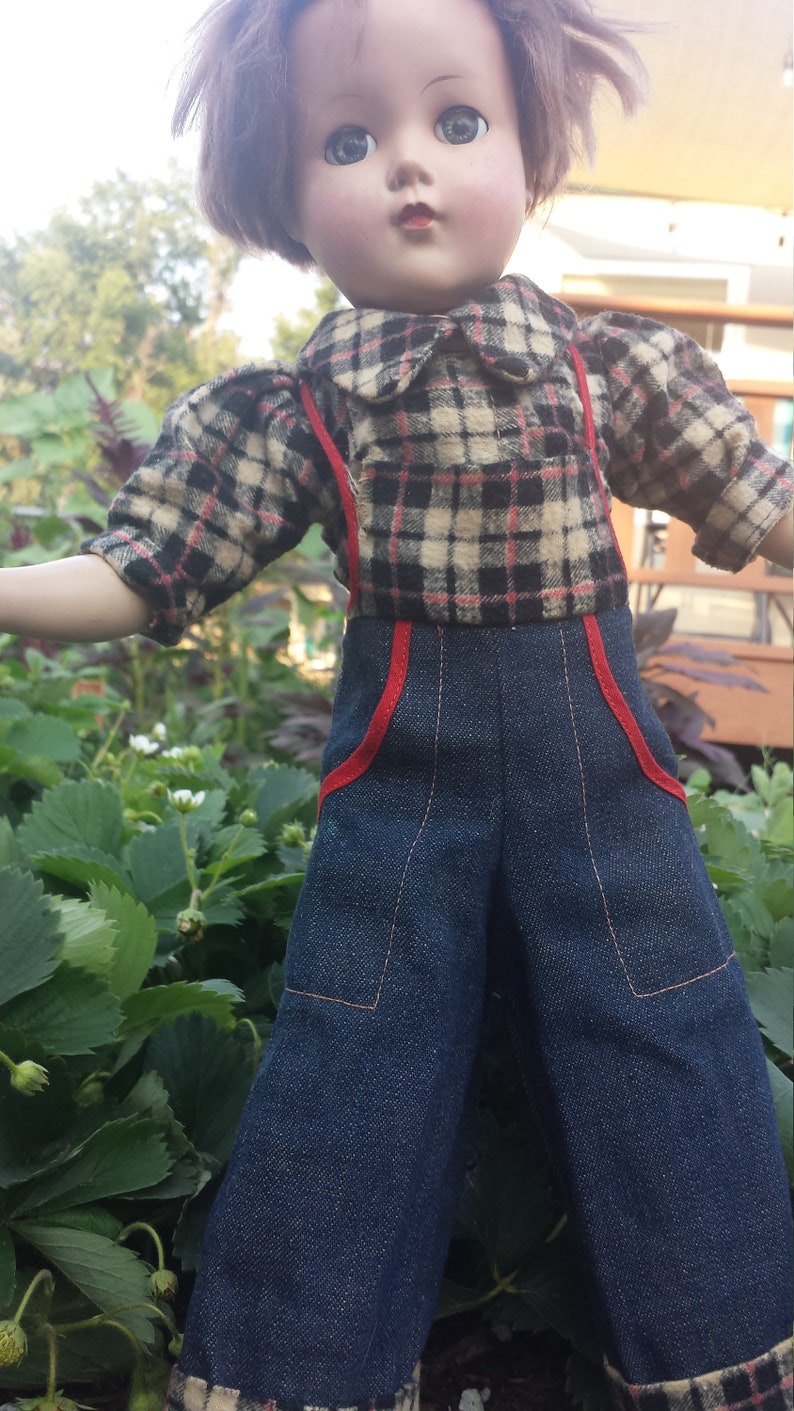 May include: A vintage doll with brown hair, wearing a plaid shirt and blue denim overalls with red trim. The doll is standing in a garden setting.
