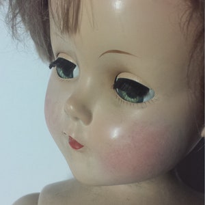 May include: A vintage porcelain doll with brown hair, green eyes, and a painted red mouth. The doll has a realistic face and is wearing a simple white dress.