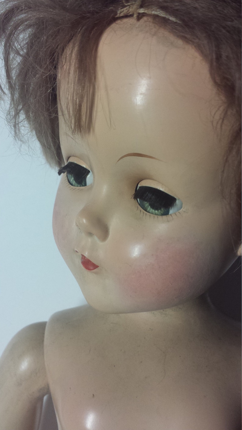 May include: A vintage porcelain doll with brown hair, green eyes, and a painted red mouth. The doll has a realistic face and is wearing a simple white dress.