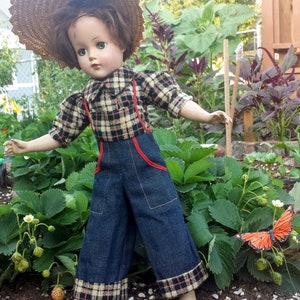 May include: A vintage doll wearing a plaid shirt, denim overalls, and a straw hat. The doll is standing in a garden with green plants and a butterfly.