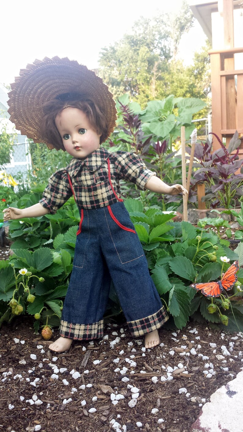 May include: A vintage doll wearing a plaid shirt, denim overalls, and a straw hat. The doll is standing in a garden with green plants and a butterfly.
