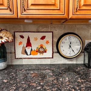 Pode incluir: Um pendurado de parede acolchoado bege com um gnomo, uma abóbora e um peru. O pendurado de parede está em uma bancada de cozinha com um relógio e uma lanterna.