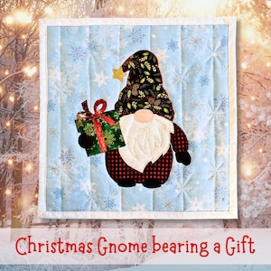 May include: A Christmas-themed quilted wall hanging featuring a gnome holding a gift. The gnome has a white beard, a black and red plaid body, and a hat with a star. The gift is green with a red bow. The background is light blue with snowflakes.